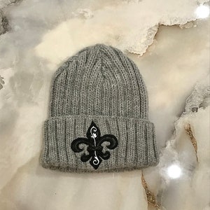 May include: Grey knitted beanie with a black embroidered fleur-de-lis design.