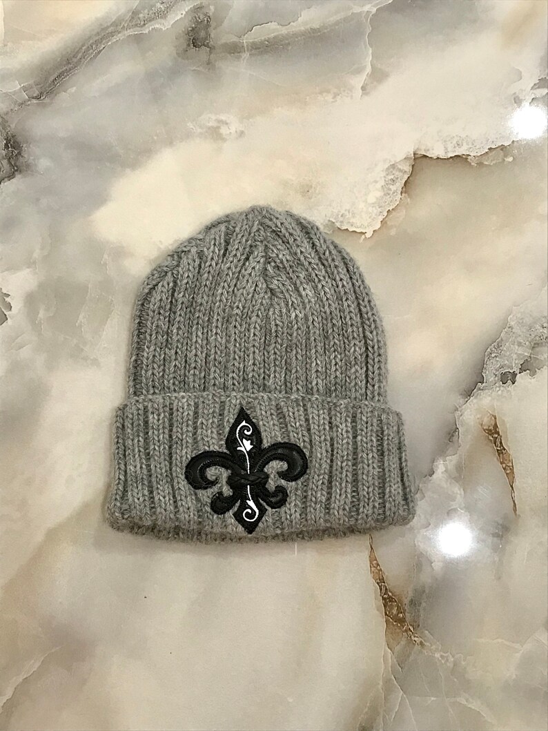 May include: Grey knitted beanie with a black embroidered fleur-de-lis design.