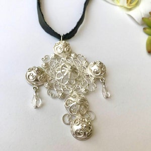 May include: A silver filigree pendant necklace with a black ribbon. The pendant is a cross shape with intricate details and clear crystal accents.