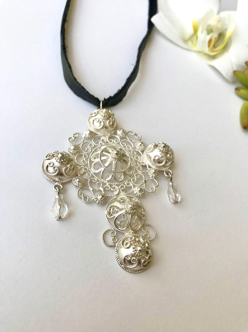 May include: A silver filigree pendant necklace with a black ribbon. The pendant is a cross shape with intricate details and clear crystal accents.