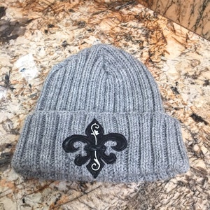 May include: Grey knitted beanie with a black and white fleur-de-lis embroidered on the front.