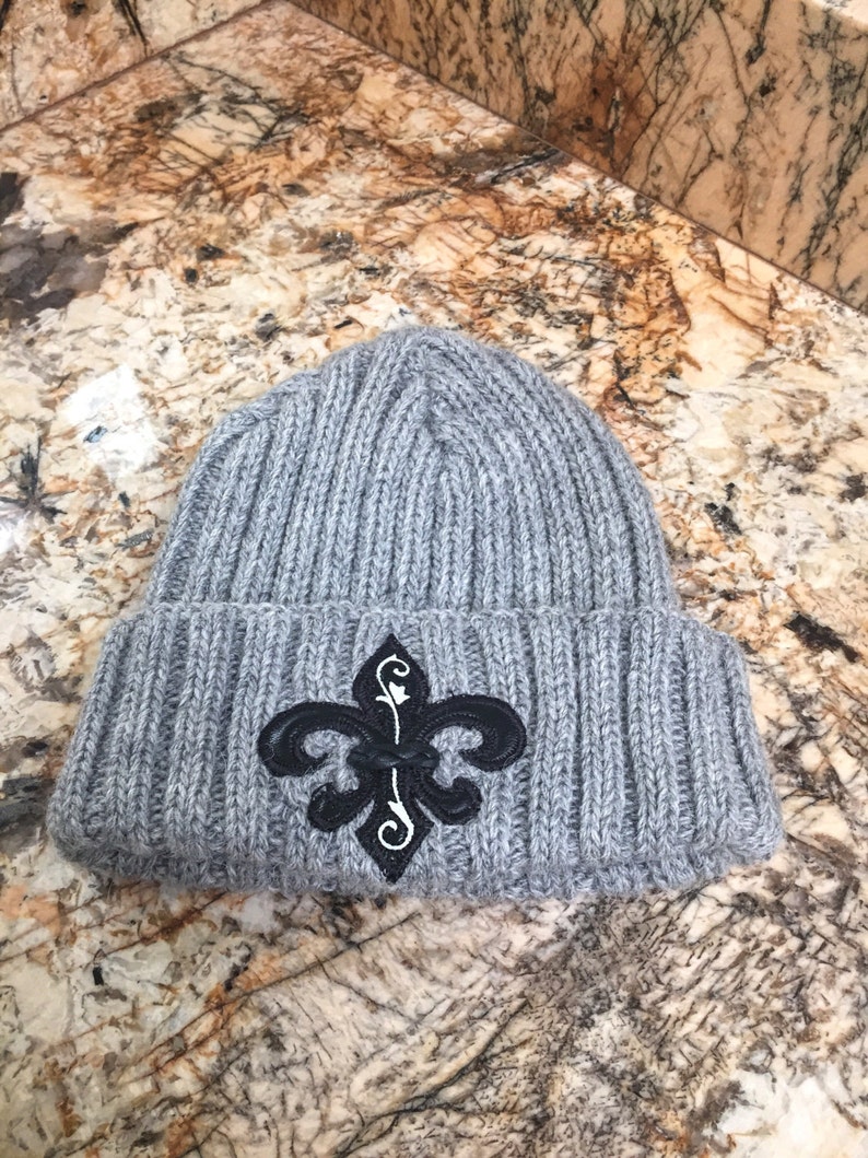 May include: Grey knitted beanie with a black and white fleur-de-lis embroidered on the front.