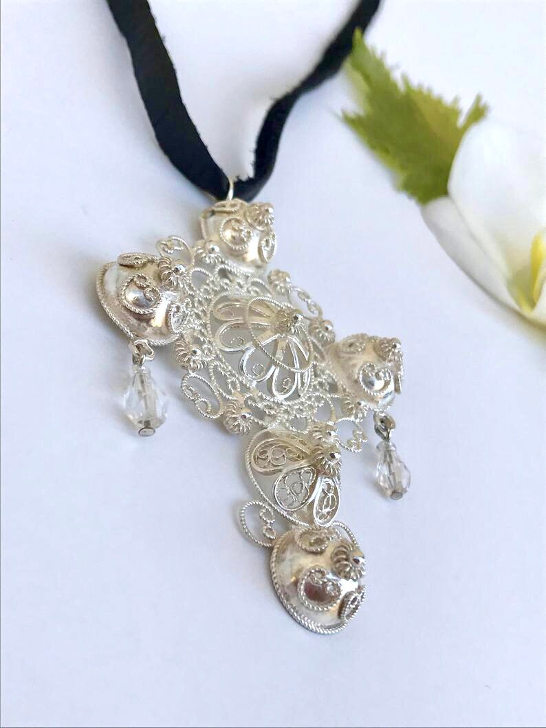 May include: A silver filigree pendant with a cross shape and crystal accents hanging from a black ribbon.