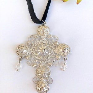 May include: A silver filigree pendant necklace with a black ribbon. The pendant features a large, intricate design with four round, ornate elements and two crystal drops.