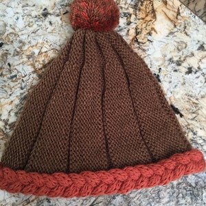 May include: A brown knitted beanie with a red braided brim and a brown and orange pom pom on top.