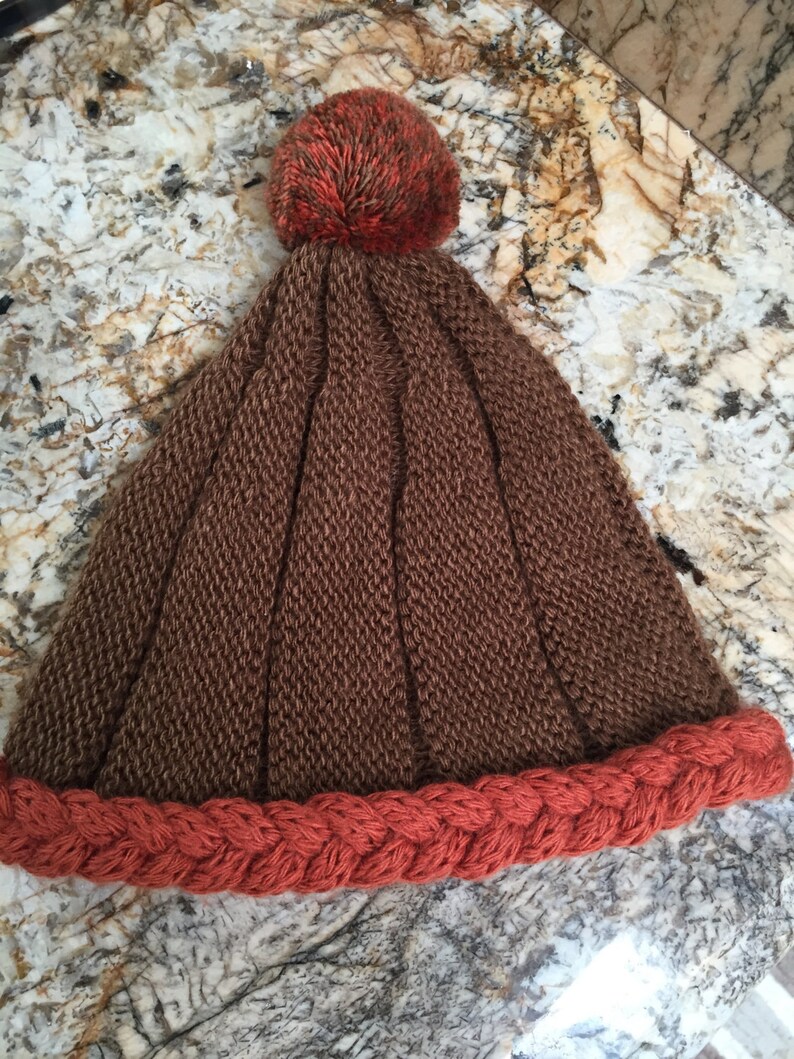 May include: A brown knitted beanie with a red braided brim and a brown and orange pom pom on top.