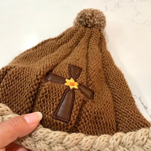 May include: Brown knitted beanie with a brown leather cross and a yellow flower embroidered on the front. The beanie has a beige braided brim and a brown pom pom on top.