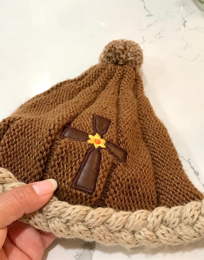 May include: Brown knitted beanie with a brown leather cross and a yellow flower embroidered on the front. The beanie has a beige braided brim and a brown pom pom on top.
