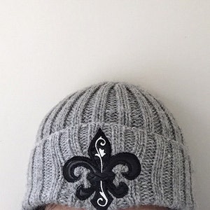 May include: Grey knitted beanie with a black and white fleur-de-lis emblem.