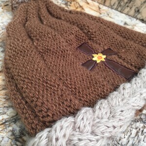 May include: Brown knitted beanie with a white pom pom and a brown cross with a yellow flower embroidered on the front. The beanie has a white braided brim.