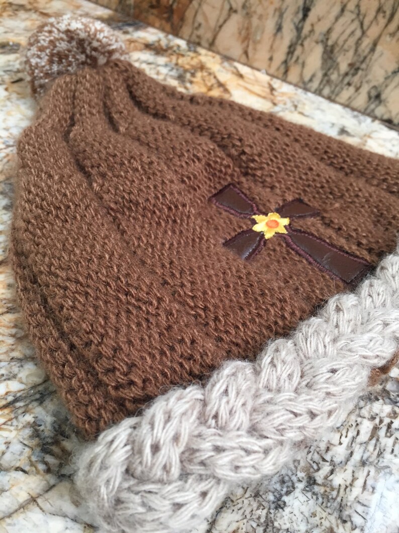 May include: Brown knitted beanie with a white pom pom and a brown cross with a yellow flower embroidered on the front. The beanie has a white braided brim.