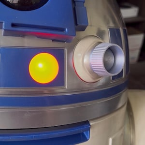 Puede incluir: Primer plano de una réplica detallada de R2-D2. El droide presenta una combinación de colores plateado y azul con una luz amarilla brillante y un componente cilíndrico. El diseño recuerda al icónico personaje de Star Wars.
