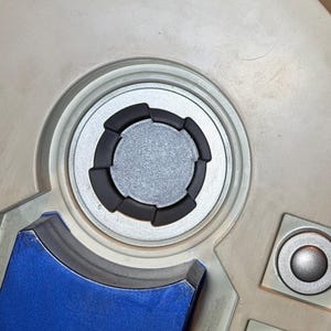 May include: Close-up of a detailed, off-white panel with a central circular design. The design features a silver center, surrounded by black segmented rings. Additional silver and blue accents are visible.