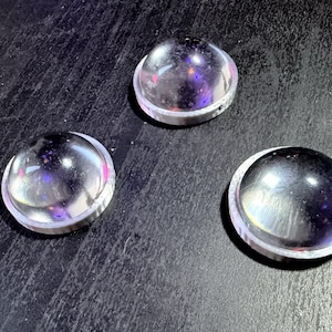 Puede incluir: Tres objetos de vidrio transparente en forma de cúpula con un borde de metal plateado. Los objetos son reflectantes, con destellos de luz púrpura y rosa. Los artículos están dispuestos sobre una superficie oscura.