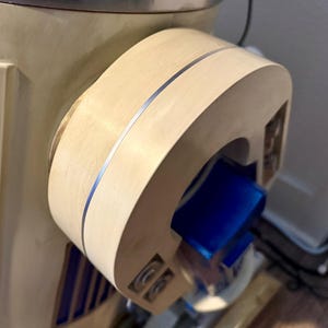 May include: A close-up shot of a light-coloured wooden component with a silver accent stripe. The component has a curved shape and a blue rectangular object is visible inside. The item appears to be part of a larger machine or appliance.