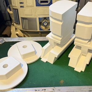 Puede incluir: Piezas de robot blancas impresas en 3D, que incluyen dos secciones grandes de la cabeza y dos piezas de base, dispuestas sobre una superficie verde. Las piezas son geométricas y angulares, con un diseño detallado. Vista parcial de un robot en el fondo.