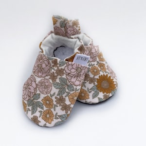 May include: A pair of baby booties with a floral print in shades of pink, brown, and yellow. The booties have a white label with the text "LOVE & CO."