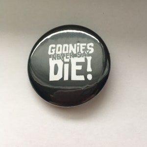 May include: A black and white button with the text "Goonies Never Say Die!" in white letters.