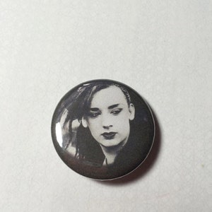 May include: A black and white button featuring a portrait of a person with dark hair and a serious expression.