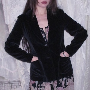 May include: A black velvet blazer with a single-button closure, worn over a floral print dress. The dress features a black background with pink and purple floral patterns. The outfit is set against a backdrop of white lace.