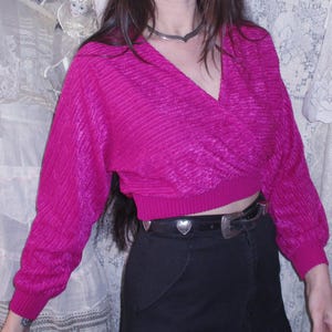 May include: A magenta wrap sweater with long sleeves and a black skirt. The sweater has a ribbed texture and a V-neck. The skirt is black and has a flared hem. A silver belt with heart-shaped details is visible.