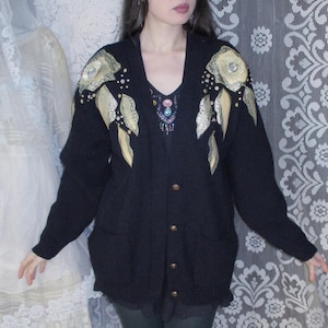 May include: A black cardigan sweater with gold and silver embellishments on the shoulders. The cardigan has a button closure and is worn over a dark green shirt and black leggings.