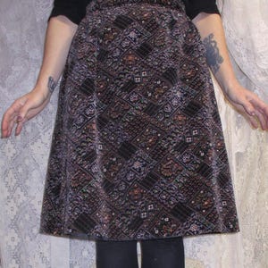 May include: A-line skirt with a geometric pattern in shades of brown, black, and rust. The skirt is paired with black leggings and knee-high lace-up boots. The outfit is set against a backdrop of white lace.