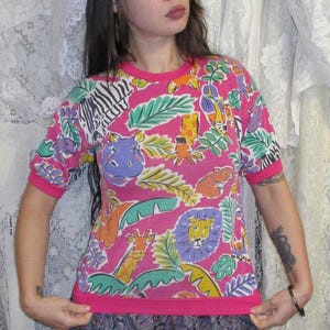 May include: A short-sleeved pink t-shirt with a vibrant jungle-themed print. The design features colorful illustrations of animals like zebras, giraffes, and lions, along with tropical leaves. The shirt has a pink trim around the neckline and sleeves.