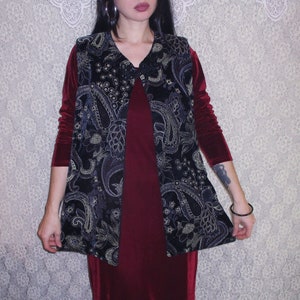 Könnte beinhalten: Eine schwarze Samt-Weste mit goldfarbenem Paisley-Muster über einem burgunderfarbenen Samtkleid.