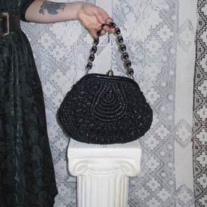 May include: A black crocheted handbag with a black chain strap. The bag is sitting on a white pedestal.