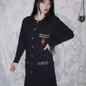 May include: A long-sleeved black dress with gold trim and buttons. The dress has a small embroidered emblem on the left chest.