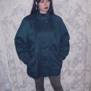 May include: A teal green jacket with a zipper and button closure. The jacket has a drawstring waist and a hood. The model is wearing the jacket with black leggings and black shoes.