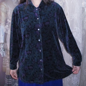 May include: A long-sleeved button-down shirt in a dark teal and purple velvet fabric featuring a tonal floral pattern. The shirt has a classic collar, a full-length front button placket with small round buttons, and a straight hemline.