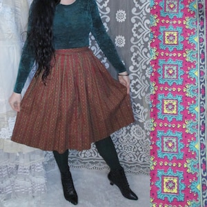 May include: A woman wearing a teal green long-sleeved shirt and a brown and gold patterned skirt. She is standing in front of a white lace curtain.