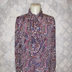 Puede incluir: Una blusa de manga larga con estampado paisley en tonos de morado, verde, dorado y marrón. La blusa tiene un cuello alto y cierre de botones en la parte delantera.