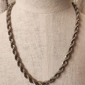 May include: Silver-toned rope chain necklace displayed on a mannequin. The necklace is made of interlocking, twisted metal links, creating a textured, rope-like appearance. The chain is draped against a neutral-colored background.