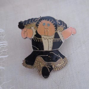 Puede incluir: Un broche de esmalte vintage que representa una figura estilizada con un atuendo negro con adornos dorados y un sombrero negro. La figura tiene una cara y brazos color melocotón, con ojos azules. El broche es probablemente de los años 80 o 90.