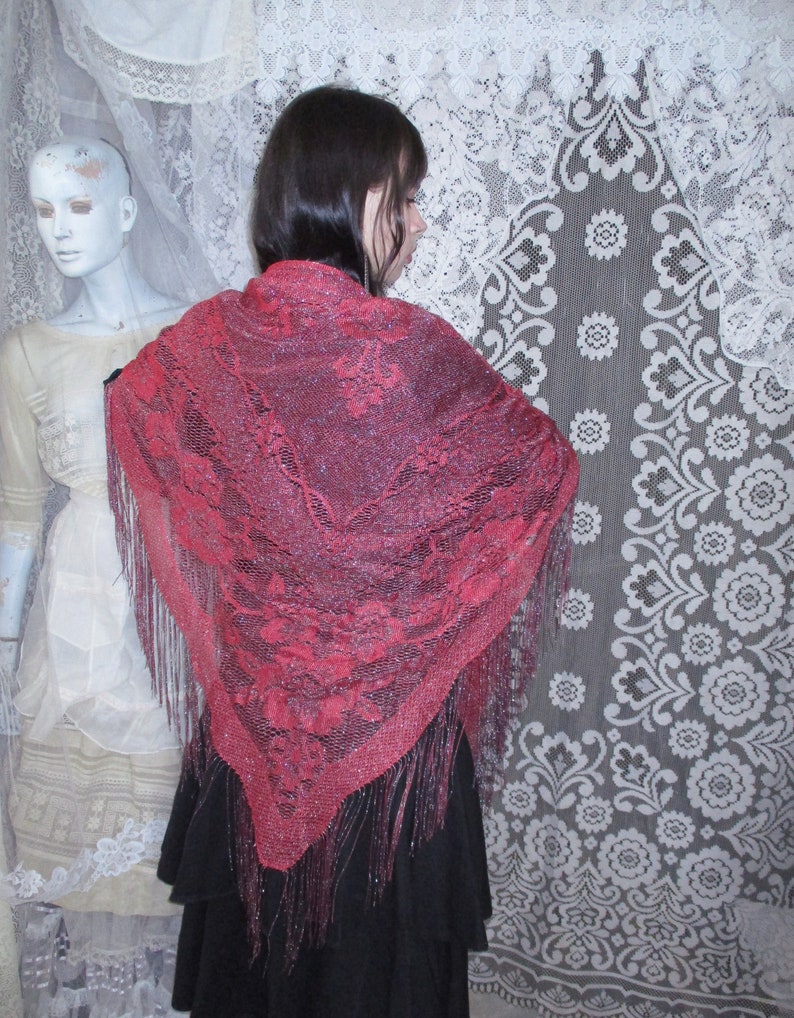 Pink Sparkly floral fringed shawl worn by a woman in all black, a long skirt and blouse, and dark hair parted to the side  with a bobby pin. A sixties mannequin wearing layers of antique white clothing is in the background, along with white lace.