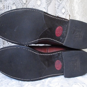 May include: Close-up of the soles of two brown leather boots. The soles are black with a textured pattern and white stitching. The heels have a red circle with text and the words "OIL RESISTANT".