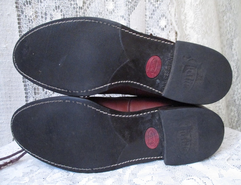 May include: Close-up of the soles of two brown leather boots. The soles are black with a textured pattern and white stitching. The heels have a red circle with text and the words "OIL RESISTANT".