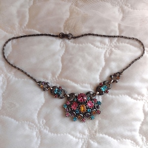 May include: A colourful necklace with a large, ornate pendant featuring a cluster of pink, blue, and orange gemstones. The pendant is set in a silver-toned metal and hangs from a delicate chain.