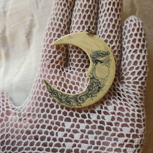 May include: Cream-colored crescent moon pendant with a detailed face design and swirling patterns in black ink. The pendant has a gold-colored loop at the top for hanging. The moon is held against a white lace background.