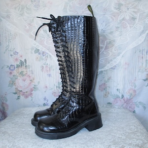 May include: Black leather knee-high boots with a crocodile embossed pattern and lace-up front. The boots have a chunky heel and a platform sole.