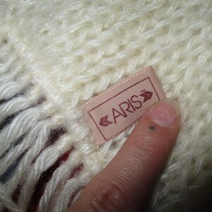 Op de afbeelding: Een witte gebreide sjaal met een roze label waarop "CARIS" in witte letters staat.