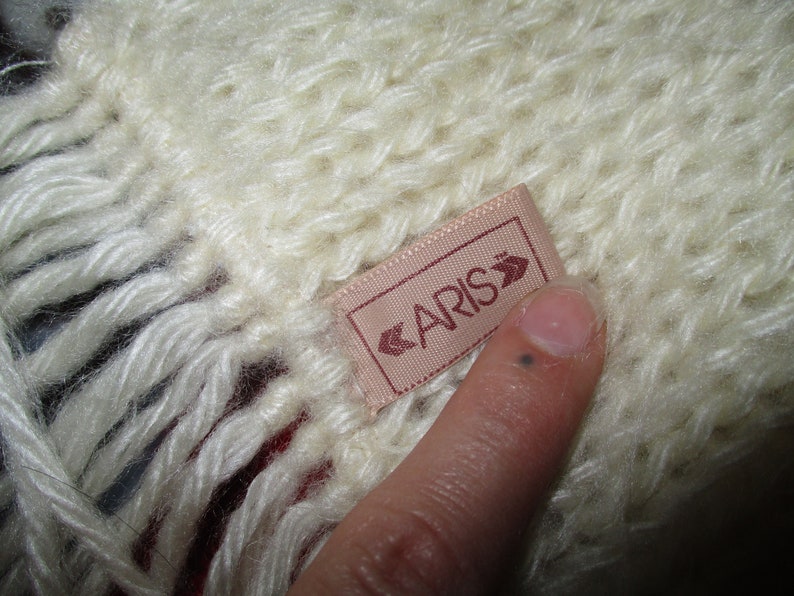 Op de afbeelding: Een witte gebreide sjaal met een roze label waarop "CARIS" in witte letters staat.