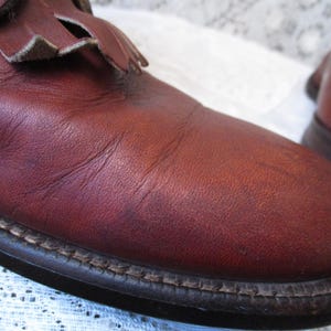 May include: Close-up of a pair of burgundy leather boots. The boots have a rounded toe, a decorative fringe detail, and a stitched sole. The leather shows natural variations in color and texture.