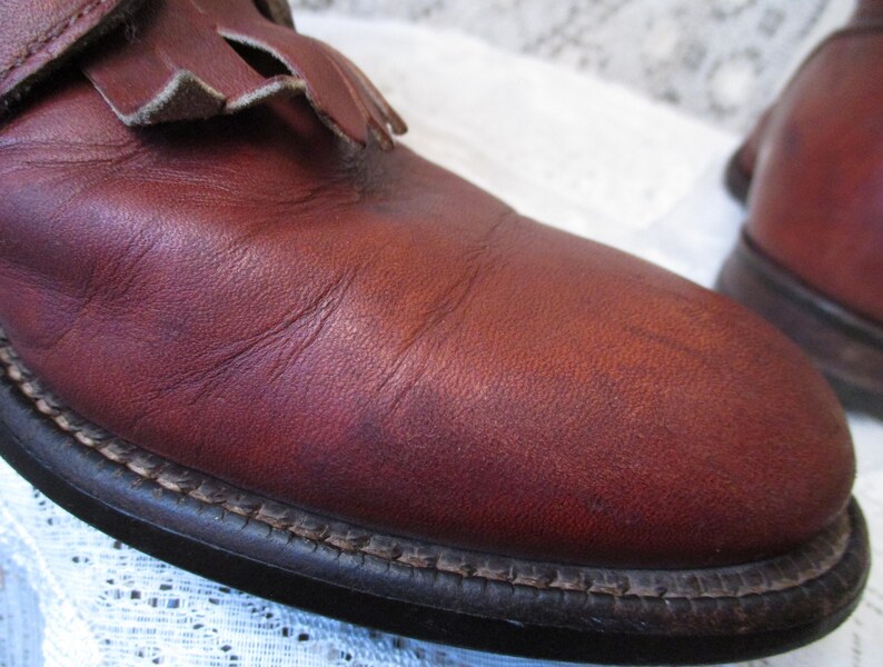 May include: Close-up of a pair of burgundy leather boots. The boots have a rounded toe, a decorative fringe detail, and a stitched sole. The leather shows natural variations in color and texture.