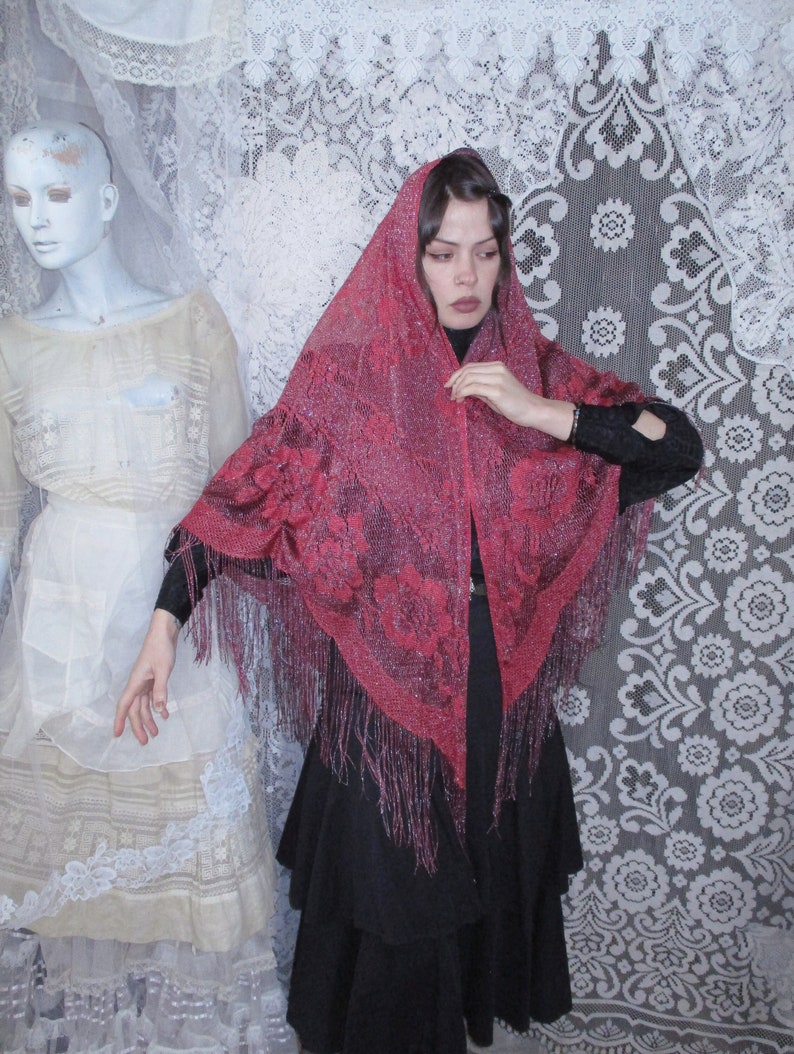 Pink Sparkly floral fringed shawl worn by a woman in all black, a long skirt and blouse, and dark hair parted to the side  with a bobby pin. A sixties mannequin wearing layers of antique white clothing is in the background, along with white lace.