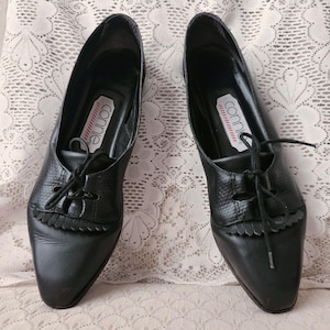 May include: A pair of black leather lace-up shoes with a textured finish on the upper section and a decorative fringe detail over the tongue. The interior insoles feature a white label with the brand name connie printed in black text.