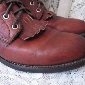 May include: Close-up of a pair of brown leather boots. The boots have a lace-up design with metal eyelets and decorative fringe on the top. The soles are dark brown with visible stitching. The leather has a worn, textured appearance.